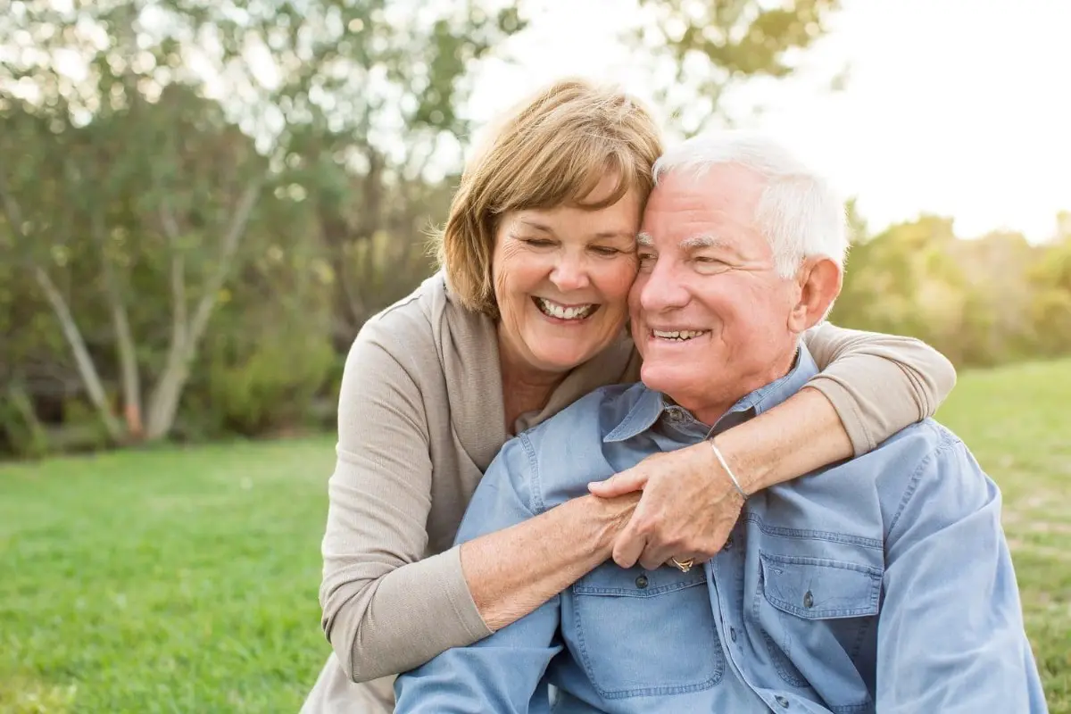 An elderly couple sits on the grass, the woman hugging the man from behind. Despite their age, they smile brightly, surrounded by greenery a testament to happiness being life's best braces or orthodontic insurance in Henderson, NV.