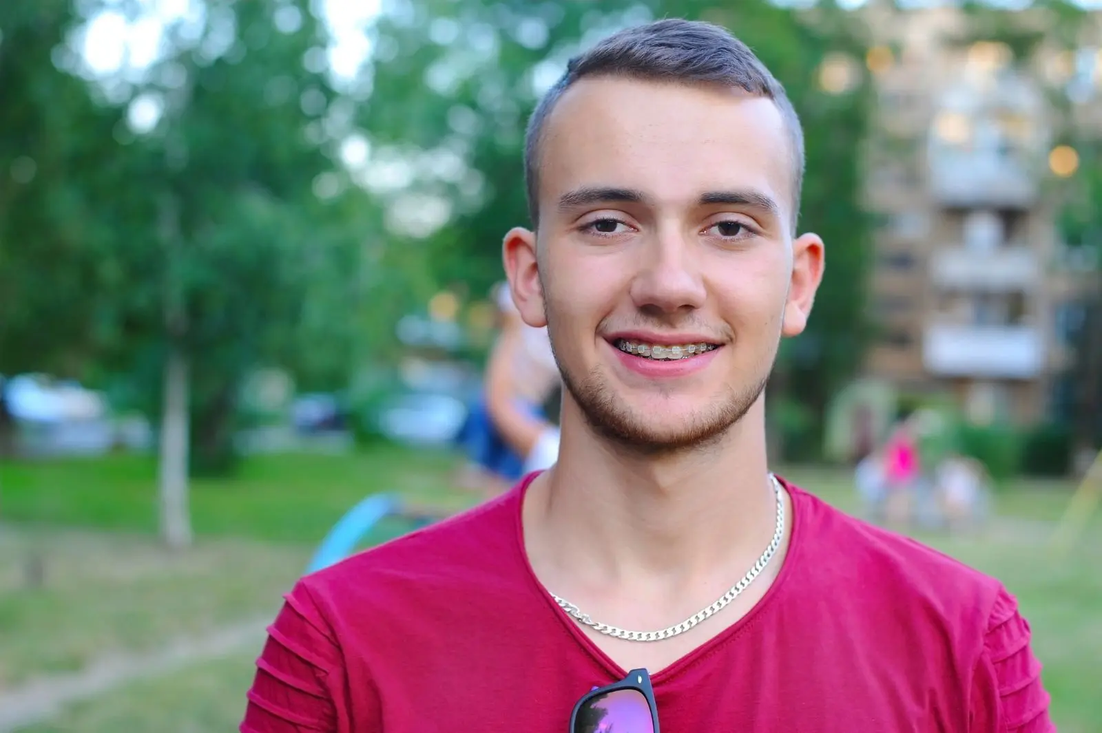 A young man with short hair and braces, wearing a red shirt and a silver chain, smiles with result clear braces from Chauncey Orthodontics in Henderson, NV.