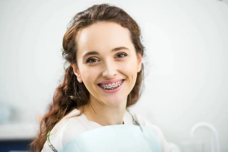 A young woman with long brown hair smiles, showing metal braces on her teeth, while seated in a dental office for change adult brace at Chauncey Orthodontics in Henderson, NV.