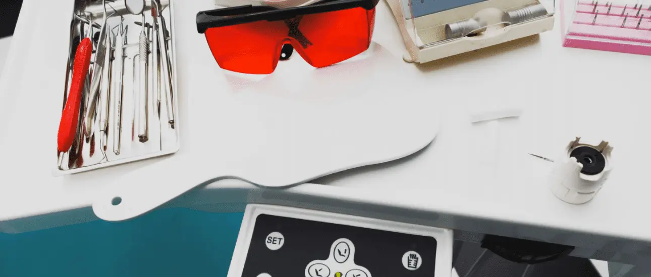 On a white surface at Chauncey Orthodontics in Henderson NV, dental tray tools including metal braces instruments, a handheld mirror, red safety glasses and equipment controls are displayed.