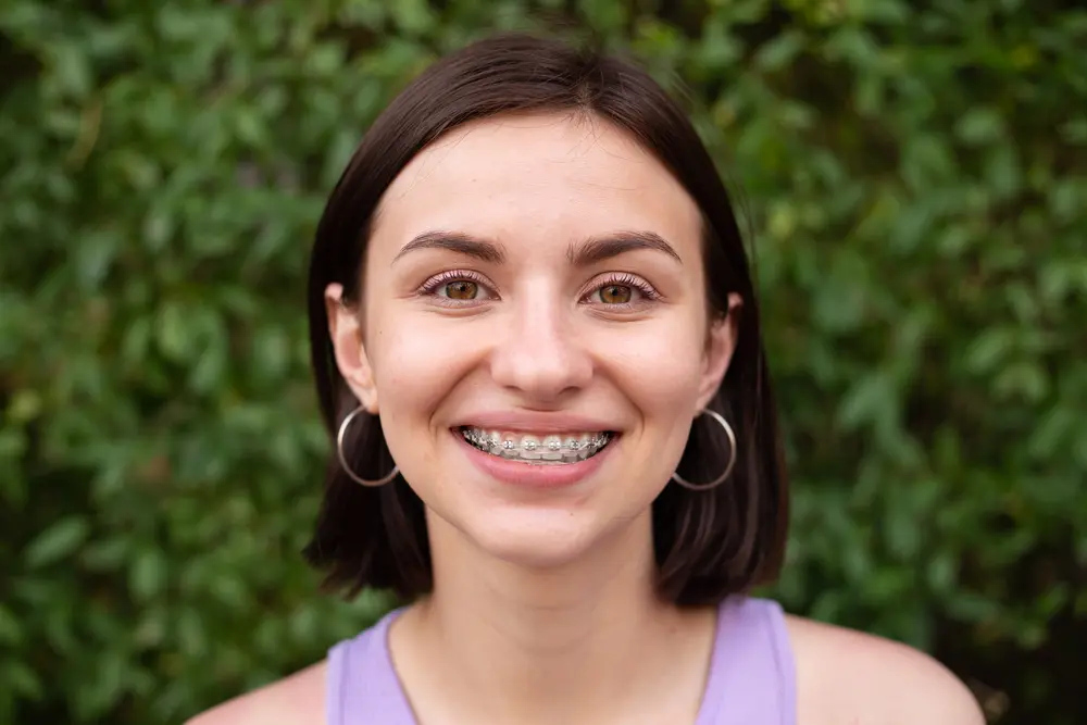 A young woman with short brown hair, wearing hoop earrings and metal braces, smiles at the camera in front of a leafy green background in Henderson, NV.