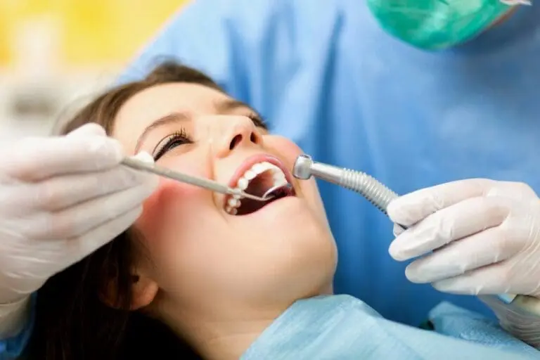A dentist wearing gloves and a mask examines a patient's open mouth using dental tools in a clinical setting.