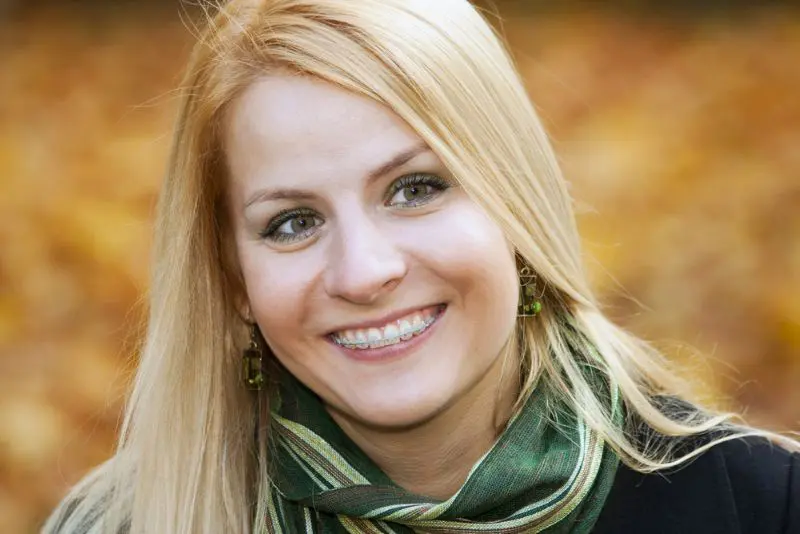 A woman with long blonde hair and showcasing clear braces smiles outdoors, wearing a green scarf and earrings, with a blurred autumn background in Henderson, NV