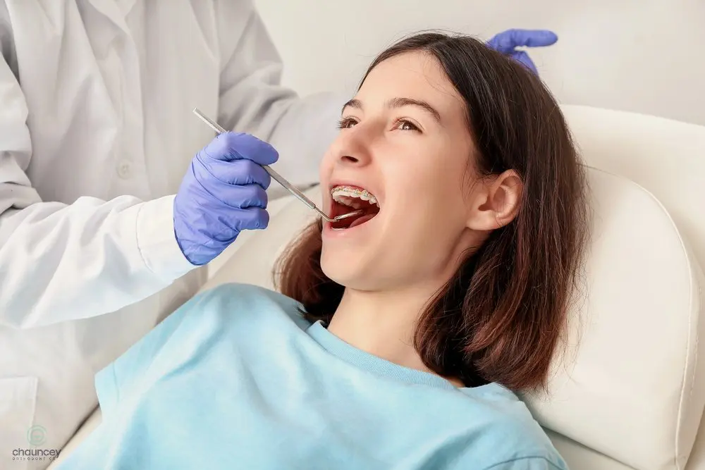 Female patient receiving a dental exam as part of her treatment plan on how to fix overbite in Henderson, NV. A dentist wearing gloves examines a teenage girl with braces, who is sitting in a dental chair with her mouth open.