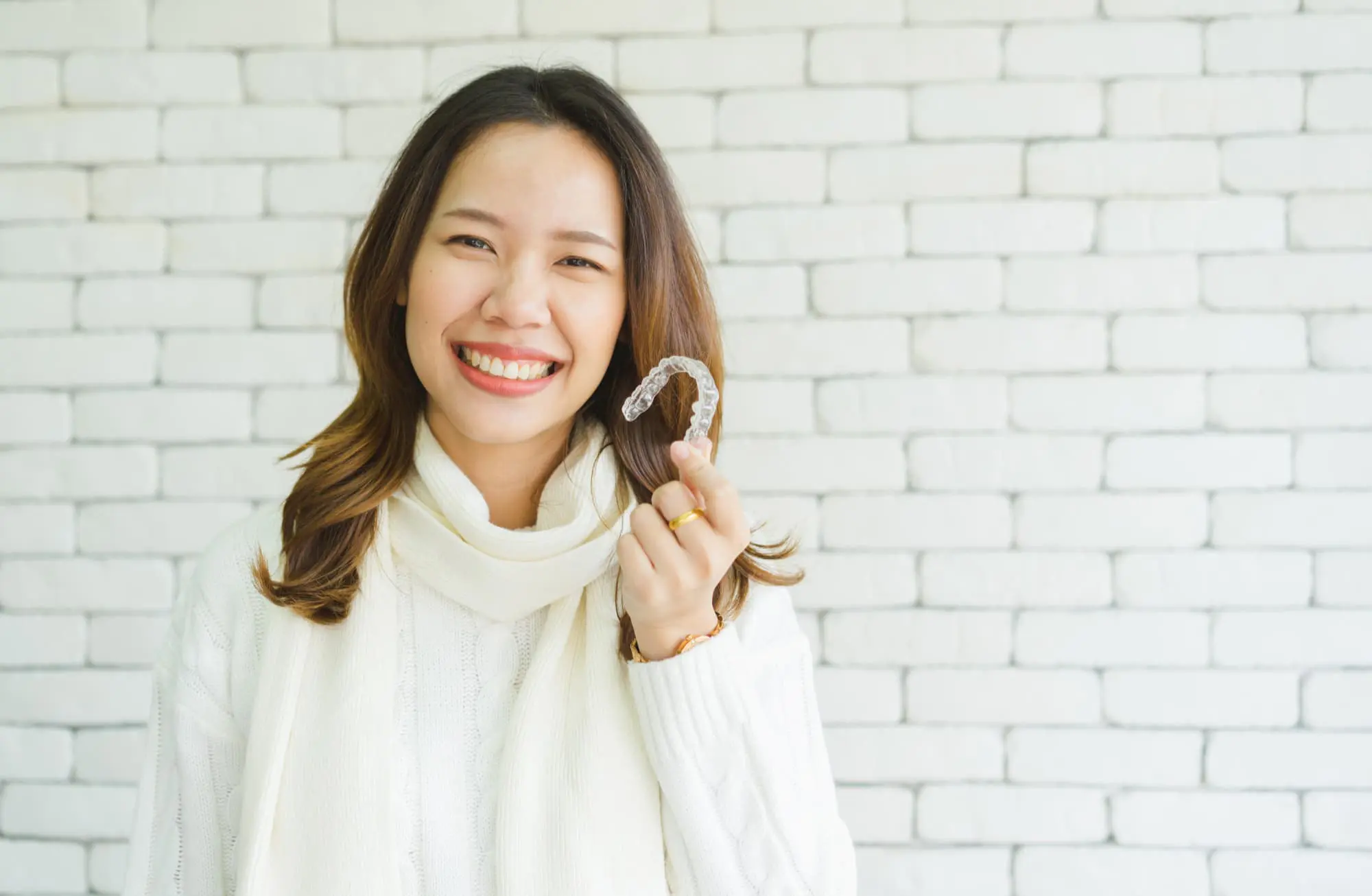 Woman in white sweater smiling and holding a clear dental aligner in front of a white brick wall.