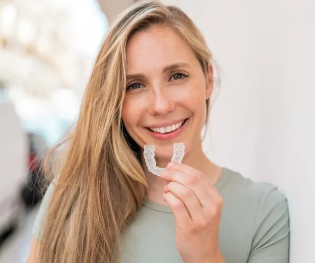 A woman smiling confidently and holding a clear dental aligner, may be benefiting from orthodontic insurance plans at Chauncey Orthodontics in Henderson, NV.