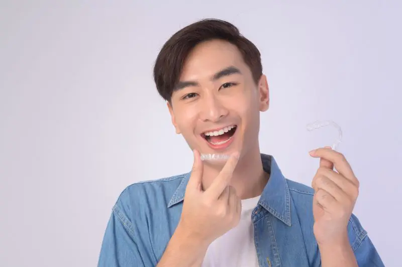 A young man wearing a denim shirt smiles while holding a Clear Aligners from invisalign, about to place it on his teeth in Henderson, NV.