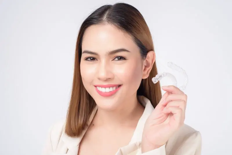 A woman with straight brown hair smiles and holds up a clear dental aligner in her right hand in Henderson, NV.