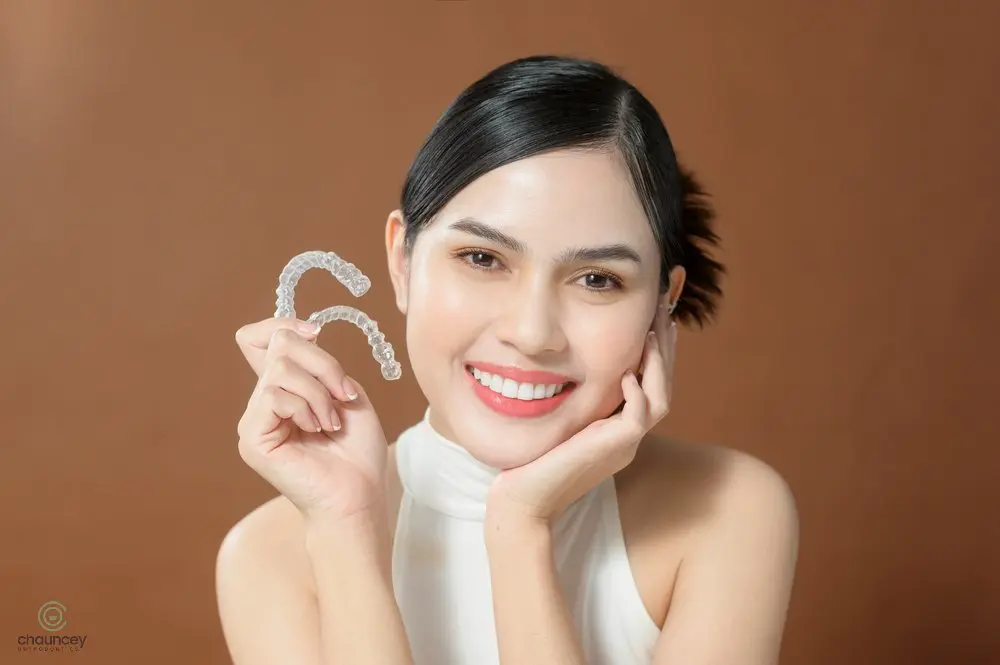 Smiling woman holding clear aligners, demonstrating how to fix overbite in Henderson, NV discreetly with Invisalign. A woman smiles while holding two clear dental aligners in her hand against a plain brown background.