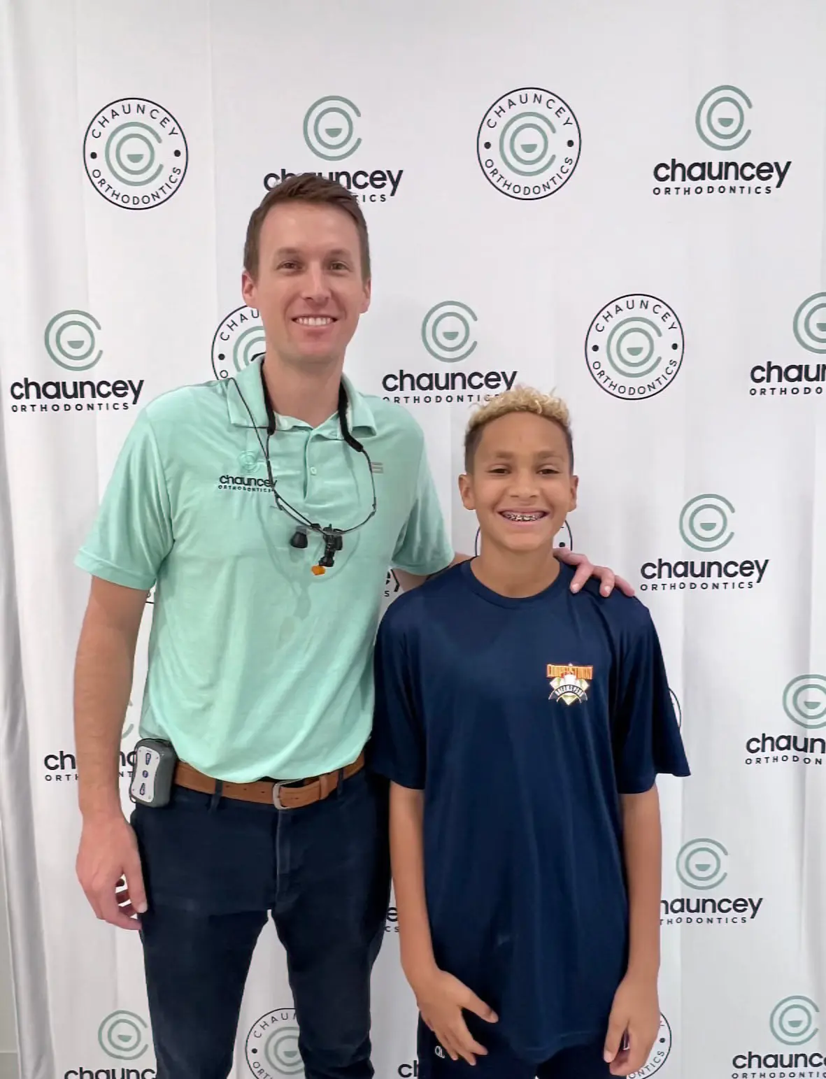 Dr. Tate Chauncey and a smiling child with braces to treat overbite stand in front of a Chauncey Orthodontics backdrop.