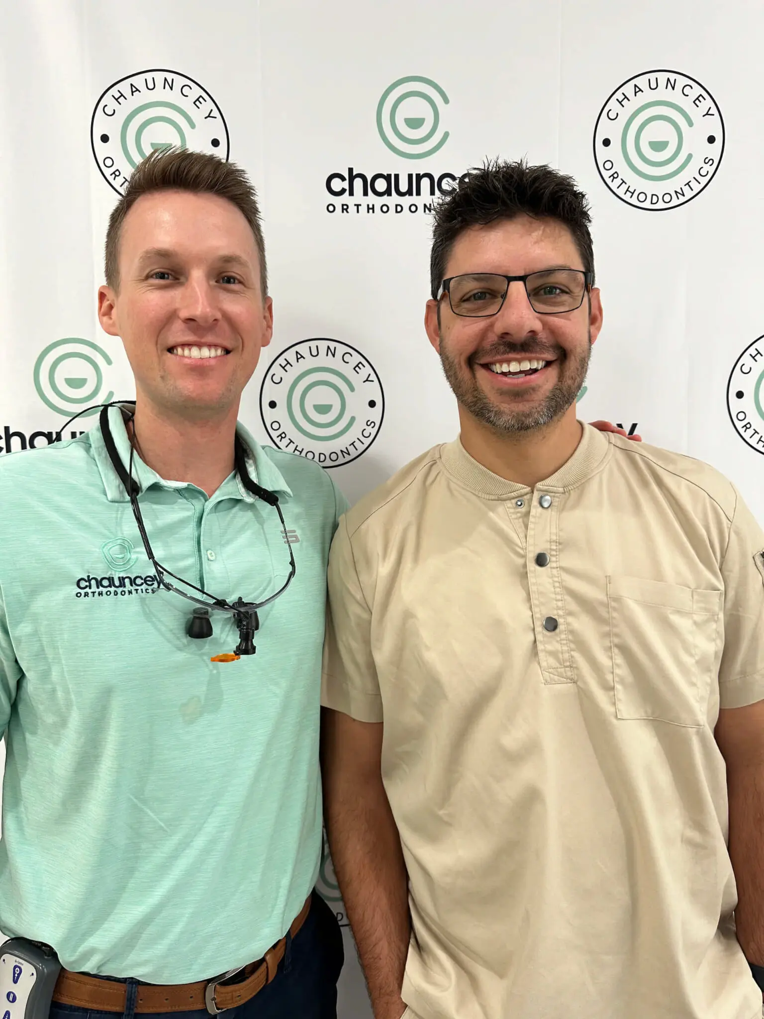 Dr. Chauncey and his patient stand side by side smiling in front of a banner that reads "Chauncey Orthodontics." One wears a mint green polo and dental loupes, the other a beige shirt.