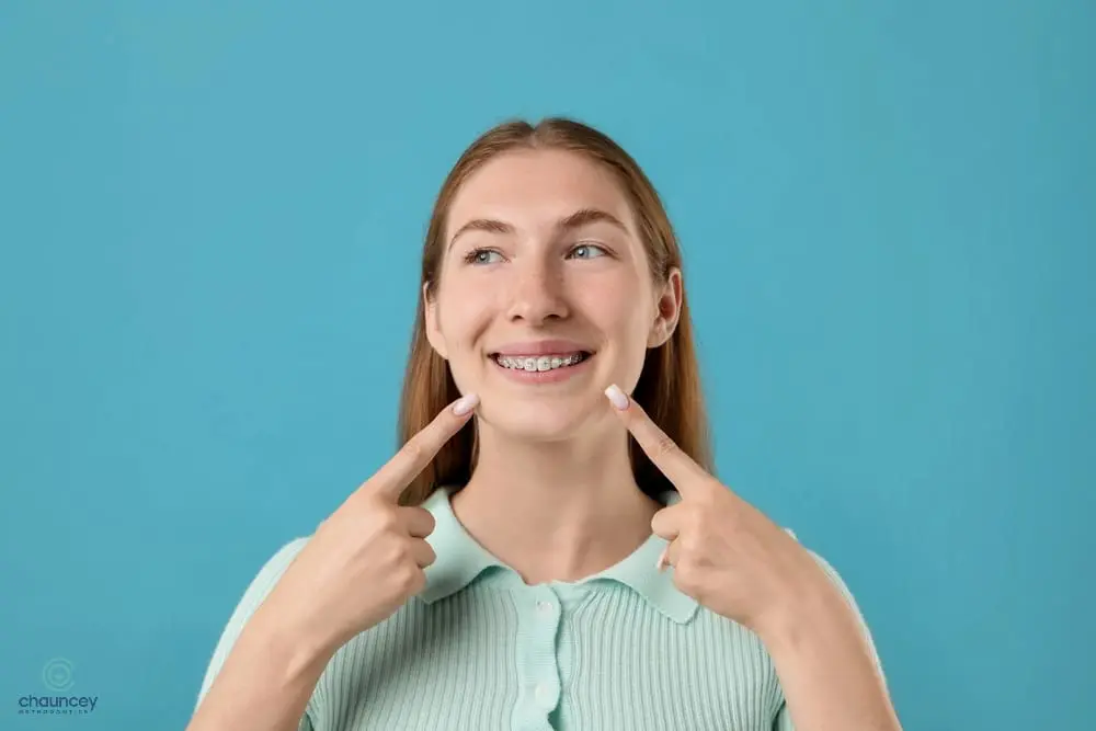 A young woman with long hair smiles and points at her teeth with braces, standing after understanding about how long do braces take in henderson, nv.