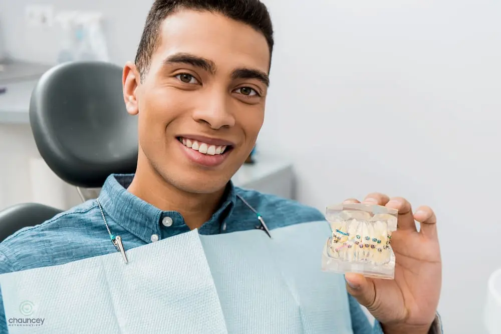 A man sitting in a dental chair smiles and holds a dental model with braces, wearing a blue shirt and a dental bib after understanding about how long do braces take in henderson, nv.