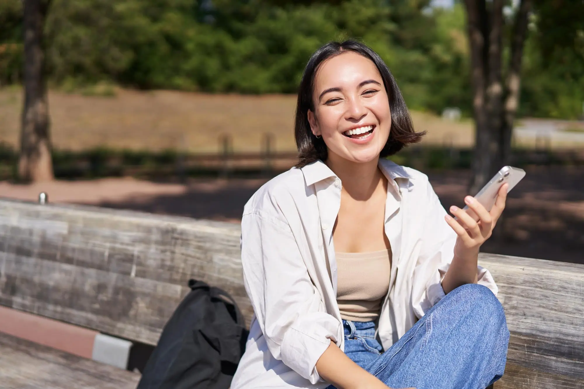 A smiling woman, who appears to be of East or Southeast Asian descent, is talking on her phone while sitting on a bench in a park, possibly in Henderson, NV.