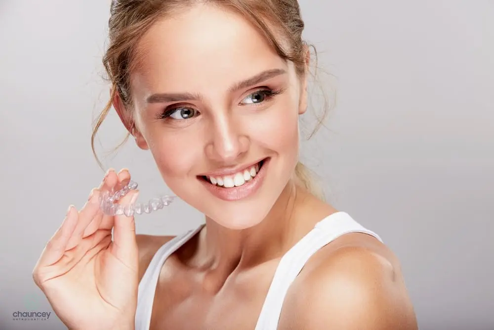 Smiling woman holding a clear dental aligner near her mouth, wearing a white tank top after understanding Does Insurance Cover Invisalign in Henderson, NV