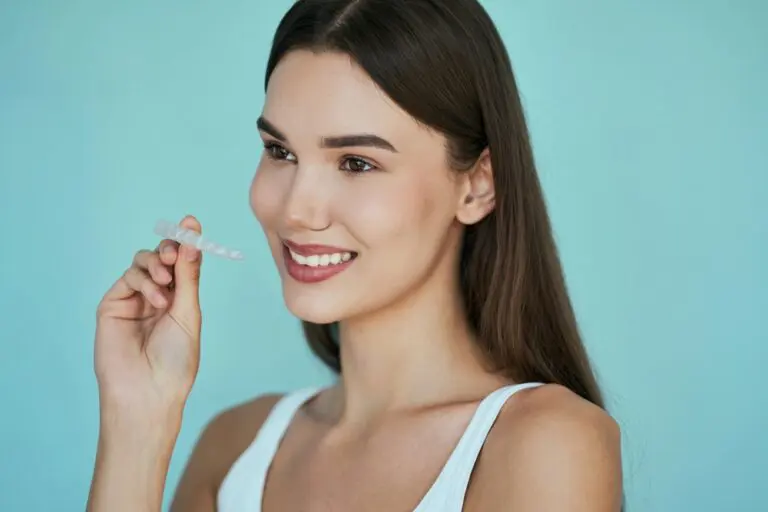 A woman with long brown hair holds a clear dental aligner near her mouth and smiles after understanding Does Insurance Cover Invisalign in Henderson, NV