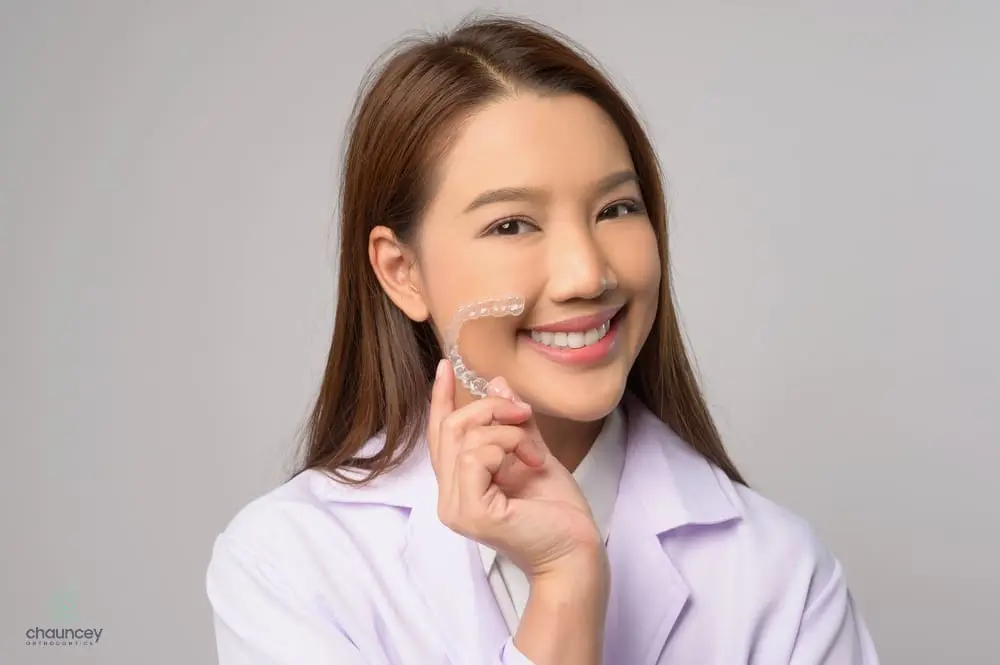 A woman in a white coat smiles and holds a clear dental aligner near her face after understanding Does Insurance Cover Invisalign in Henderson, NV