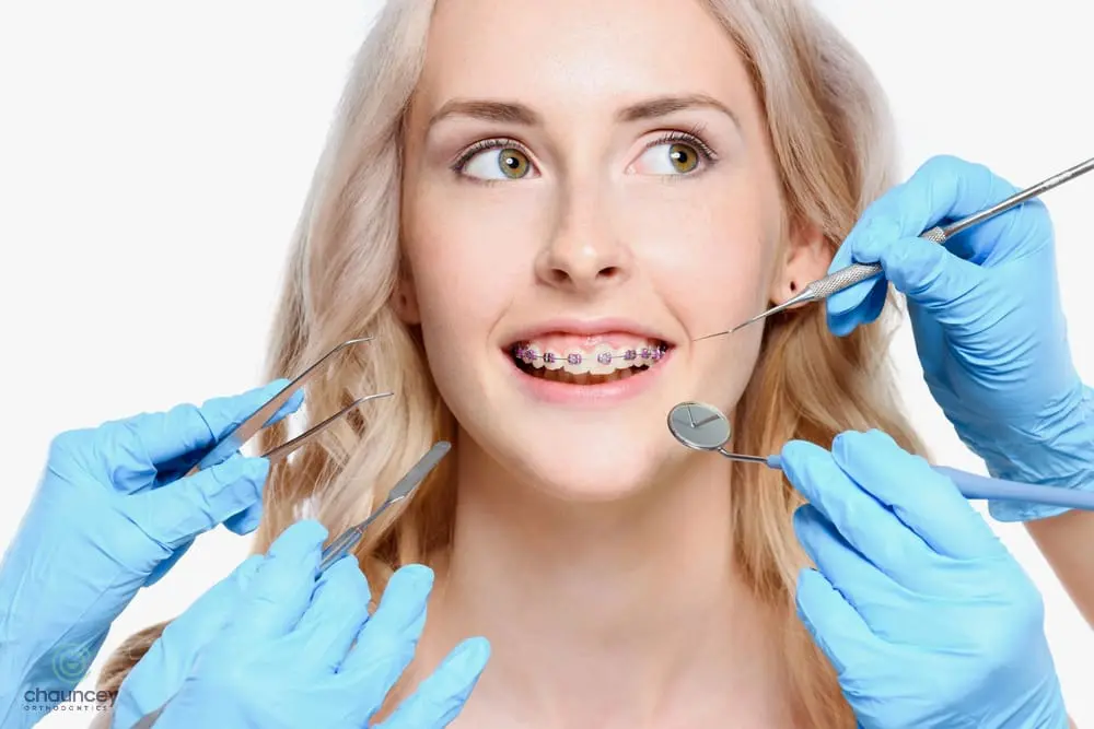 A woman with braces is examined by several gloved hands holding dental tools showcasing braces for overbite in henderson, nv.
