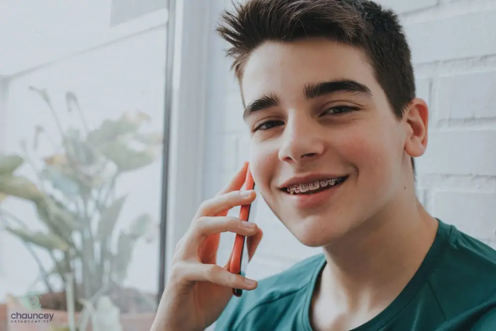 A teenage boy with braces smiles while talking on a smartphone, sitting indoors near a window with a potted plant in the background.