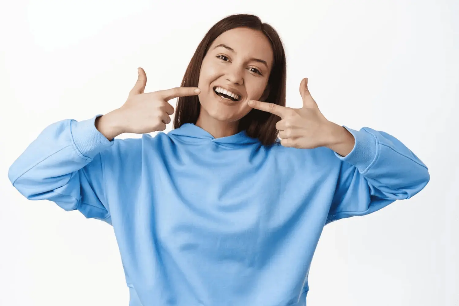 A woman in a light blue hoodie smiles and points to teeth with both index fingers, proudly displaying her results her metal braces from Chauncey Orthodontics in Henderson, NV.