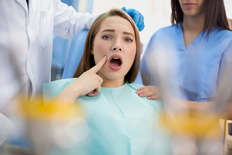 A woman sitting in a dental chair points to her mouth with a concerned expression, while two dental professionals stand beside her - Overbite vs Underbite in Henderson, NV.