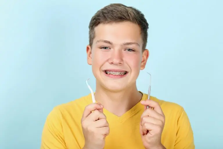 Teen boy wearing braces, smiling, and holding two dental tools in front of a light blue background - How to Floss with Braces in Henderson, NV
