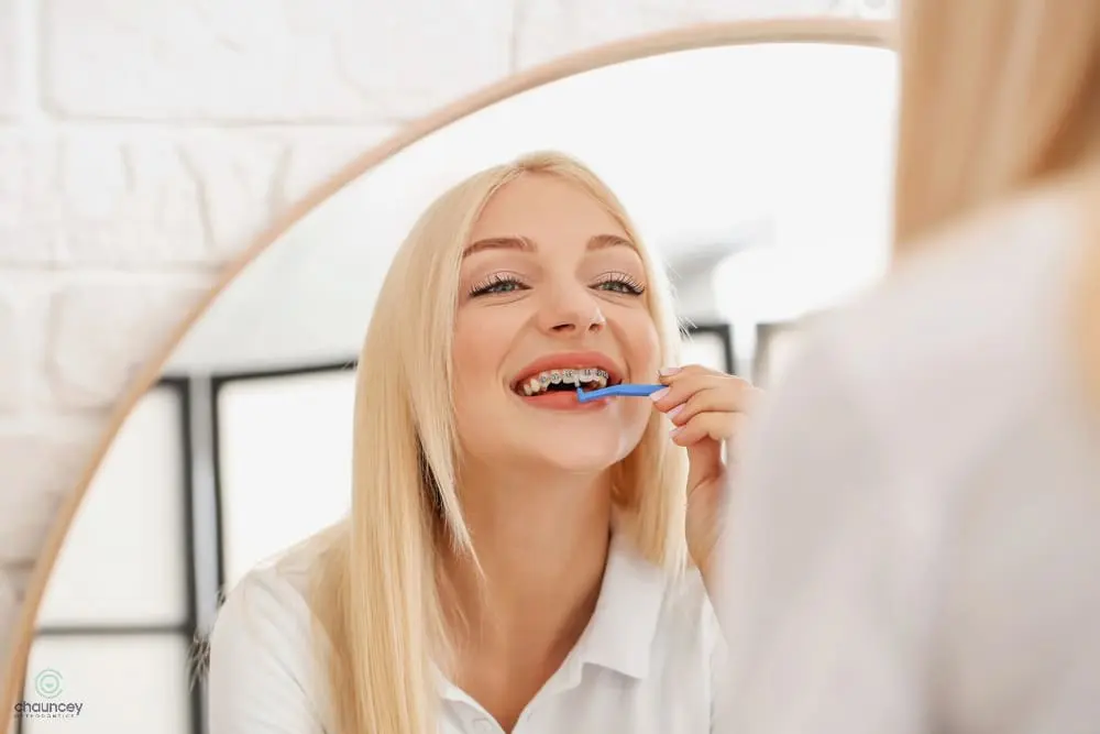 A woman with long blonde hair brushes her teeth while looking into a bathroom mirror - How to Floss with Braces in Henderson, NV