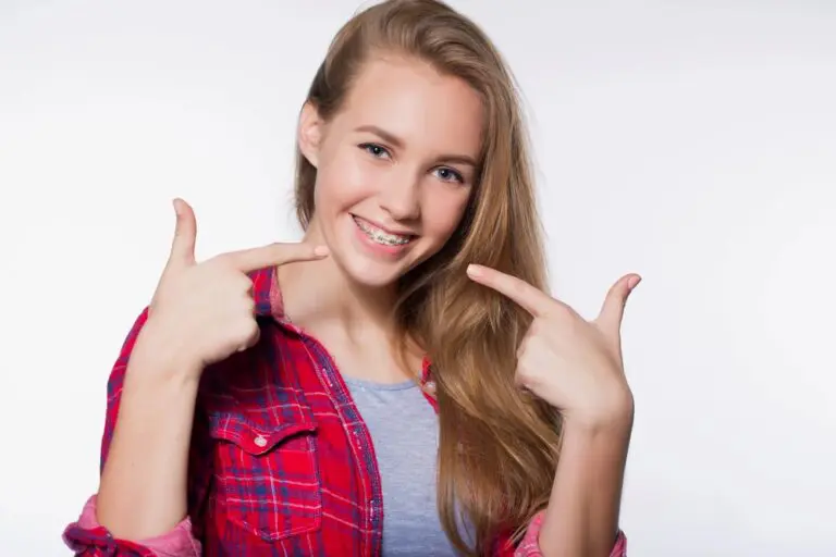 Teenage girl with long blonde hair wearing a red plaid shirt smiles and points to her braces, showing off her teeth as she thinks about how to get braces glue off teeth, against a plain white background - How to Get Braces Glue Off Teeth in Henderson, NV