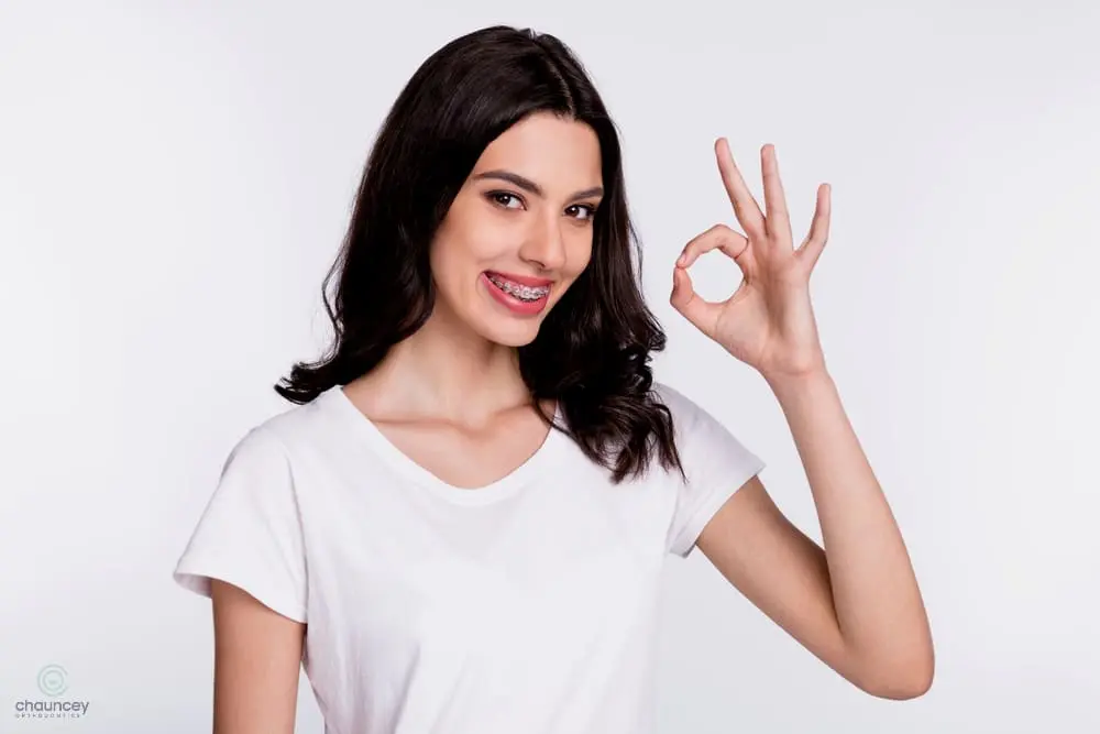 Woman with long dark hair and braces, wearing a white t-shirt, smiling and making an "OK" hand gesture against a plain light background - How to Get Braces Glue Off Teeth in Henderson, NV