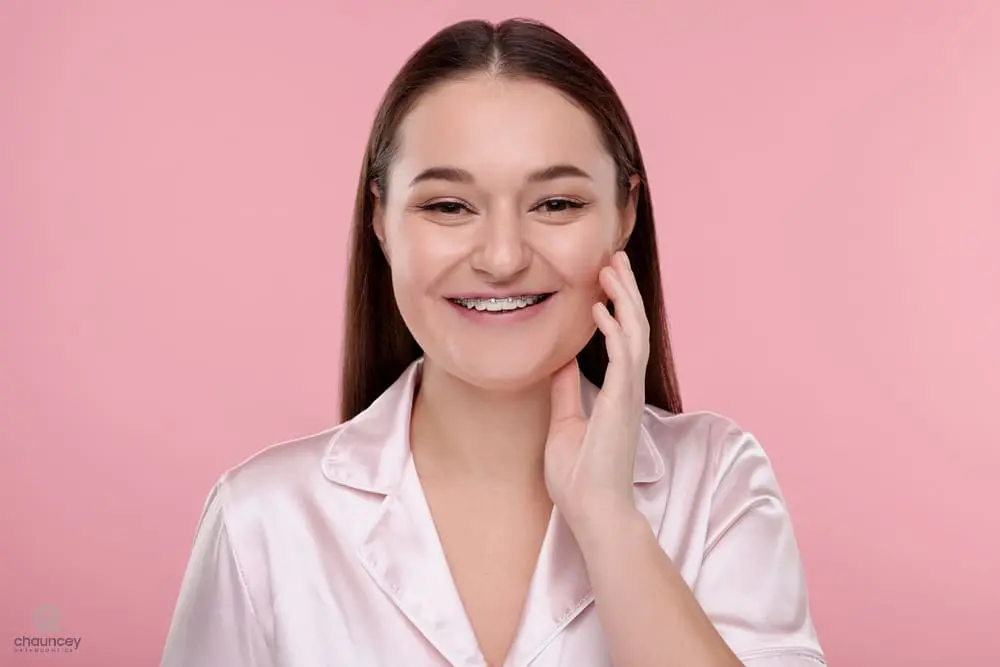 A young woman with long brown hair and braces smiles, touching her cheek with one hand, wearing a light pink satin shirt against a plain pink background - How to Get Braces Glue Off Teeth in Henderson, NV 