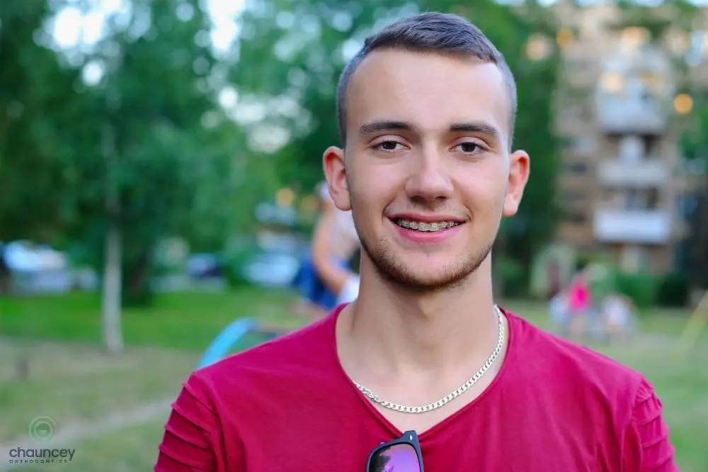 A young man with short hair and braces, wearing a red shirt and silver chain, smiles outdoors in a park-like setting - Alternatives to Braces in Henderson, NV