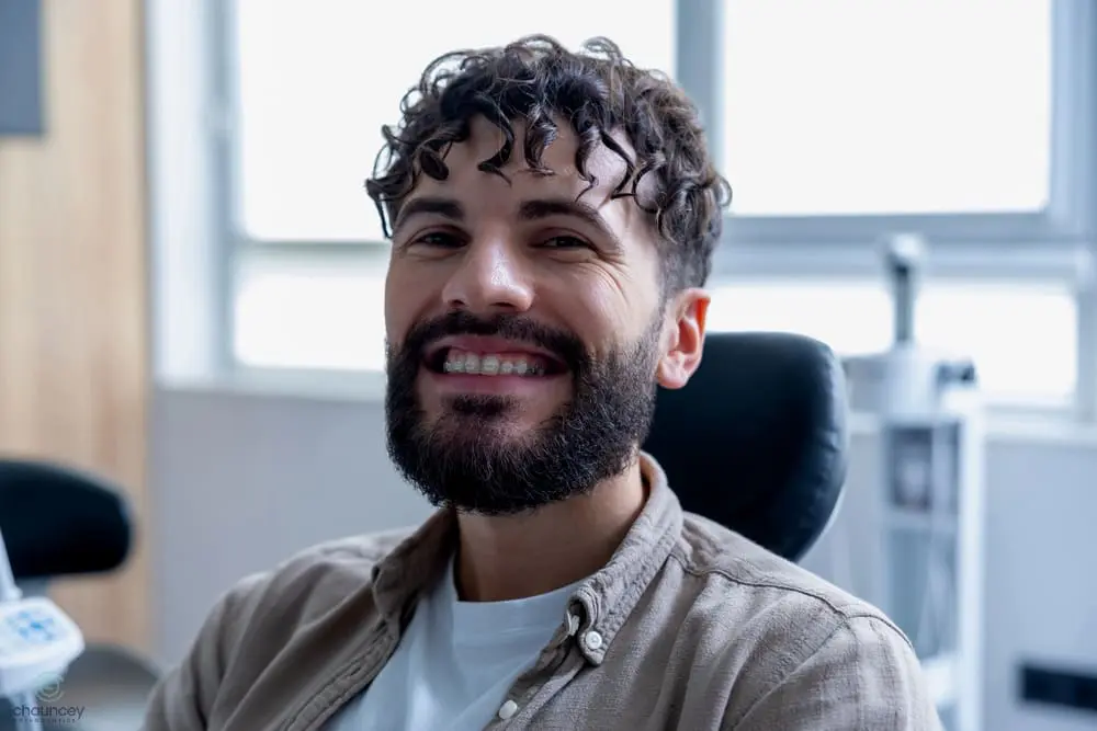 A man with curly hair and a beard is sitting in a chair, smiling, with braces on his teeth. The background appears to be a dental or medical office - Clear Braces vs Metal Braces in Henderson, NV 