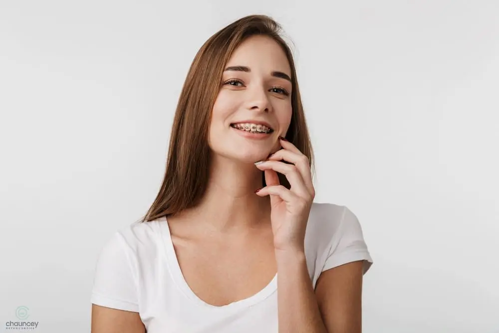 Young woman with long brown hair wearing a white t-shirt, smiling and showing braces on her teeth, against a plain light background - Clear Braces vs Metal Braces in Henderson, NV 