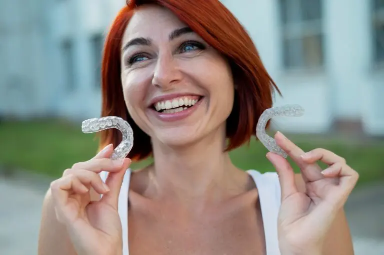 Smiling person with red hair holds two clear dental aligners in both hands outdoors - Aligners vs Braces in Henderson, NV