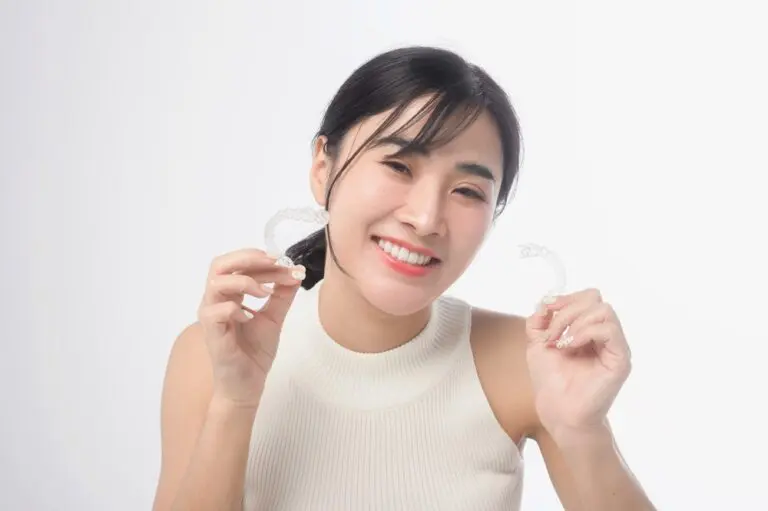 A woman smiles while holding two clear dental aligners, one in each hand, against a plain white background - How Does Invisalign Work in Henderson, NV