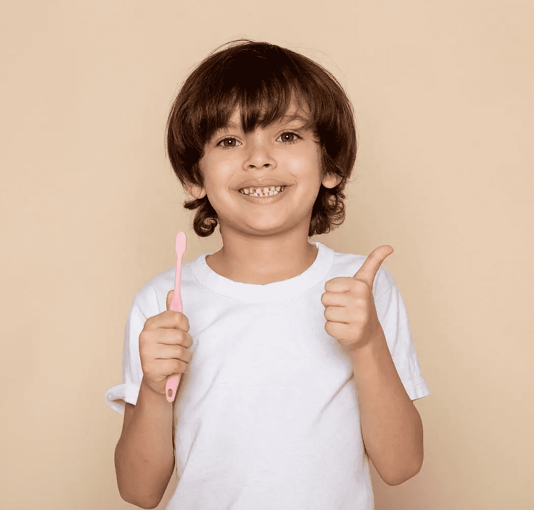 A young child in a white t-shirt, with braces and a bright smile, holds a pink toothbrush and gives a thumbs-up after visiting children orthodontist at Chauncey Orthodontics in Henderson, NV.