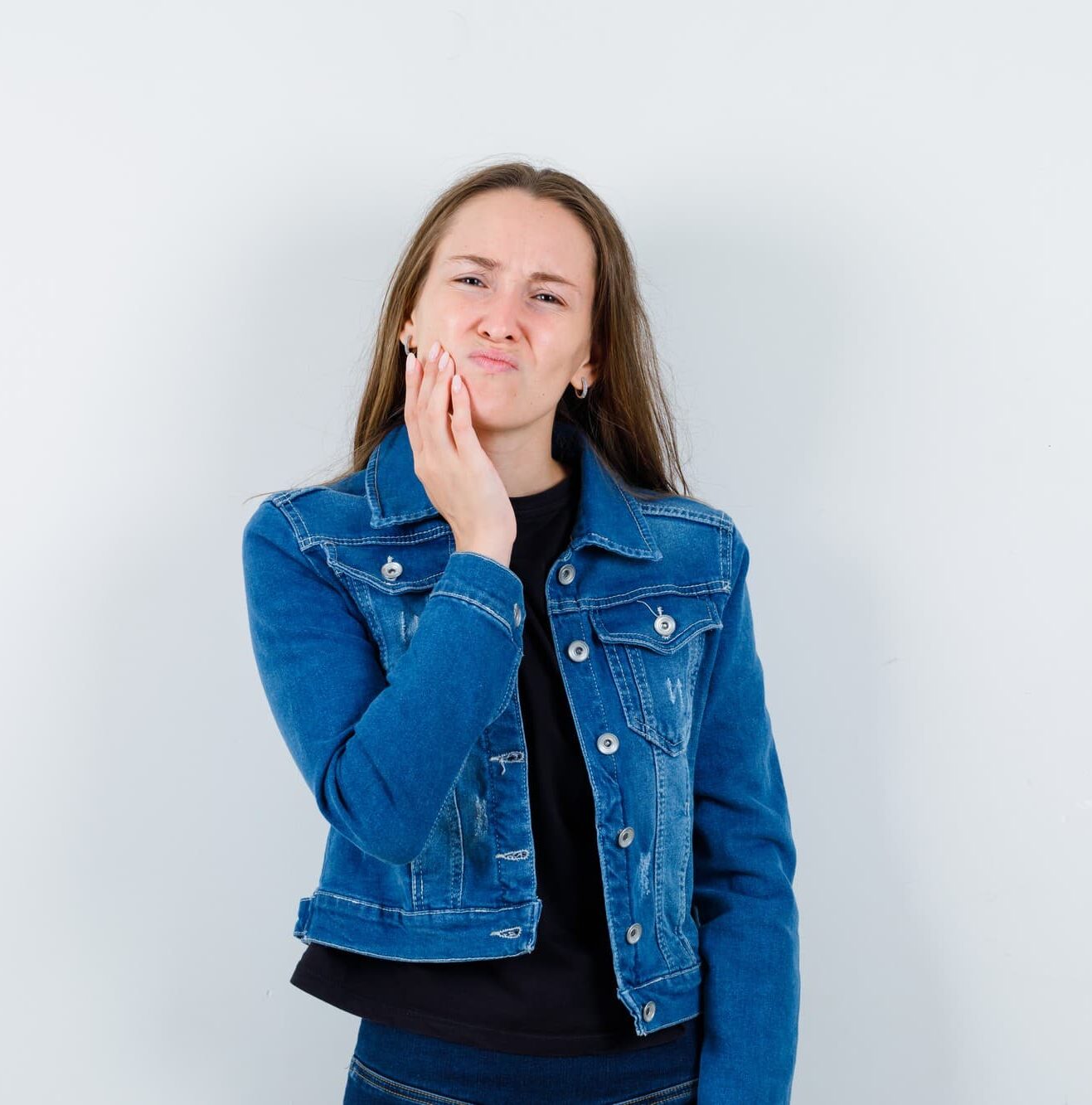 A woman in a denim jacket touches her cheek and looks uncomfortable, possibly experiencing underbite tooth pain, standing at Chauncey Orthodontics in Henderson, NV.