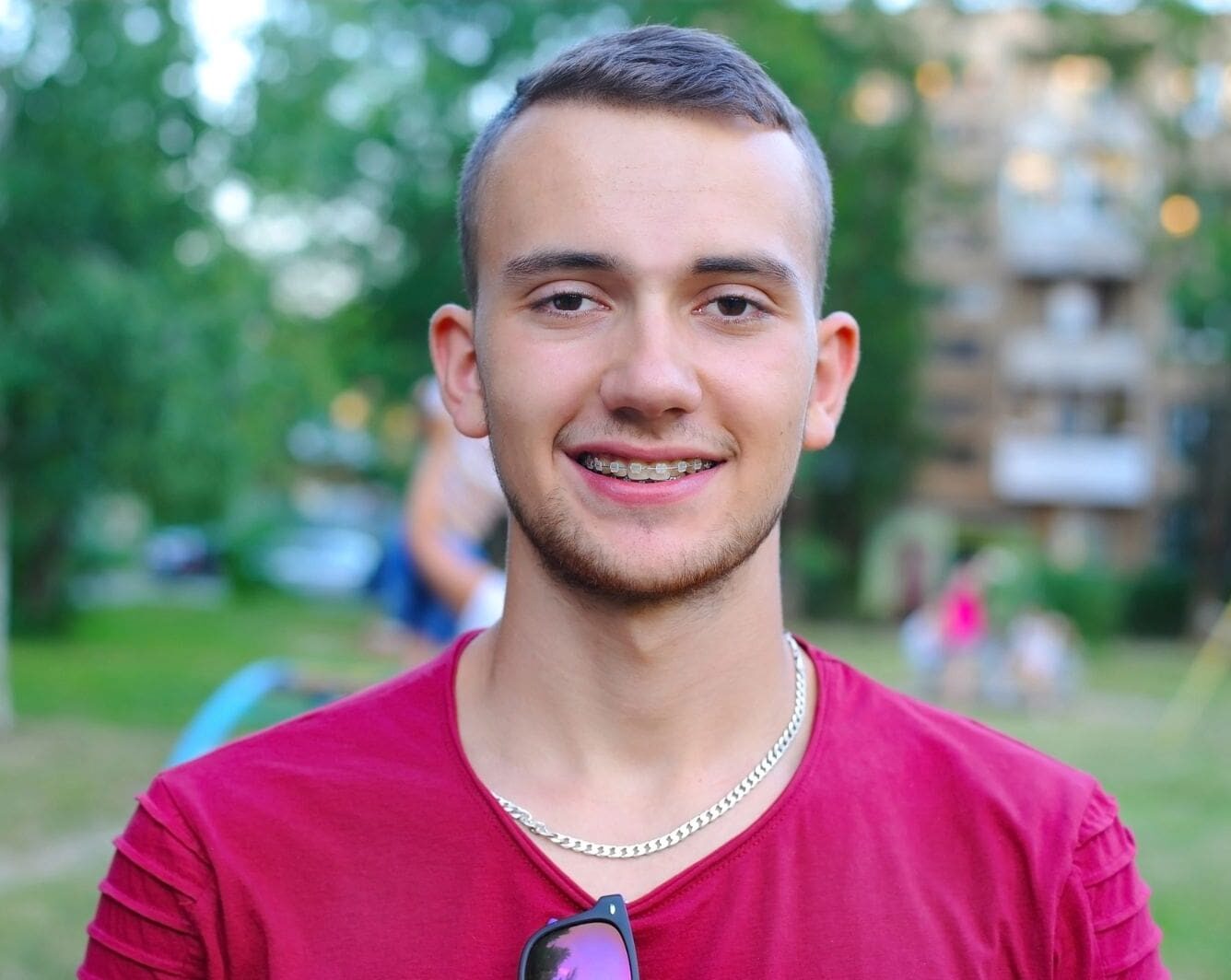 A young man with short hair and clear braces, wearing a red shirt and a silver chain, smiles outdoors with greenery and buildings in the background.